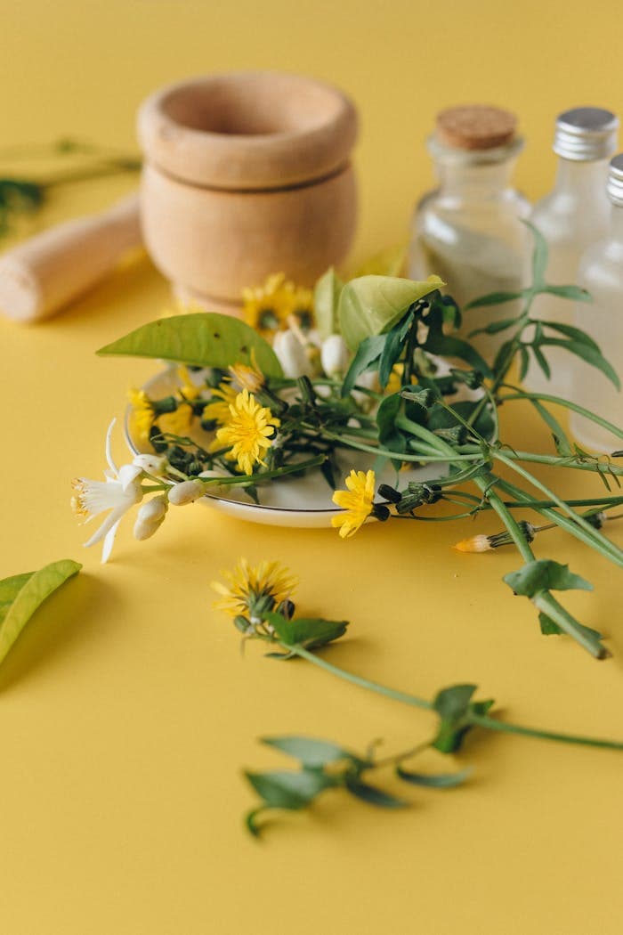 Vibrant herbal and flower arrangement with glass bottles and mortar on a yellow backdrop.